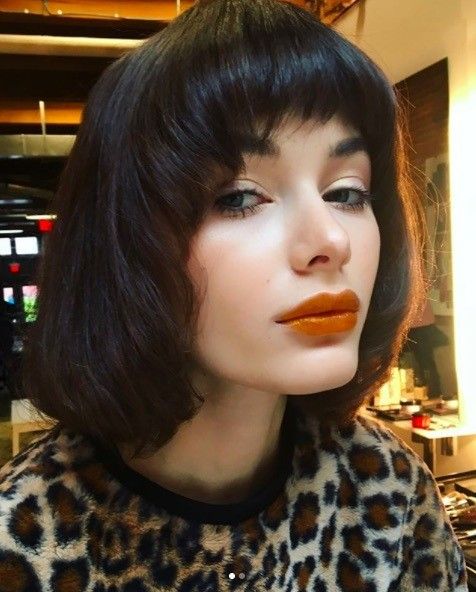 a woman with short brown hair, bangs, and orange lipstick looks at the camera. she is wearing a brown and white leopard print sweater.  she's up close and you can see her clear skin, freckles, and long eyelashes.  she's in a professional makeup setting with a makeup counter in the background.