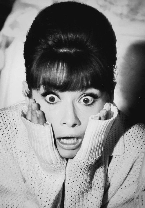 a black and white portrait of a woman with wide eyes and an open mouth, appearing surprised or scared. her hair is styled in a beehive, and she is wearing a sweater. she looks straight at the camera and holds her hands up to her face, as if in shock. the image is vintage and appears to be taken in a studio setting.