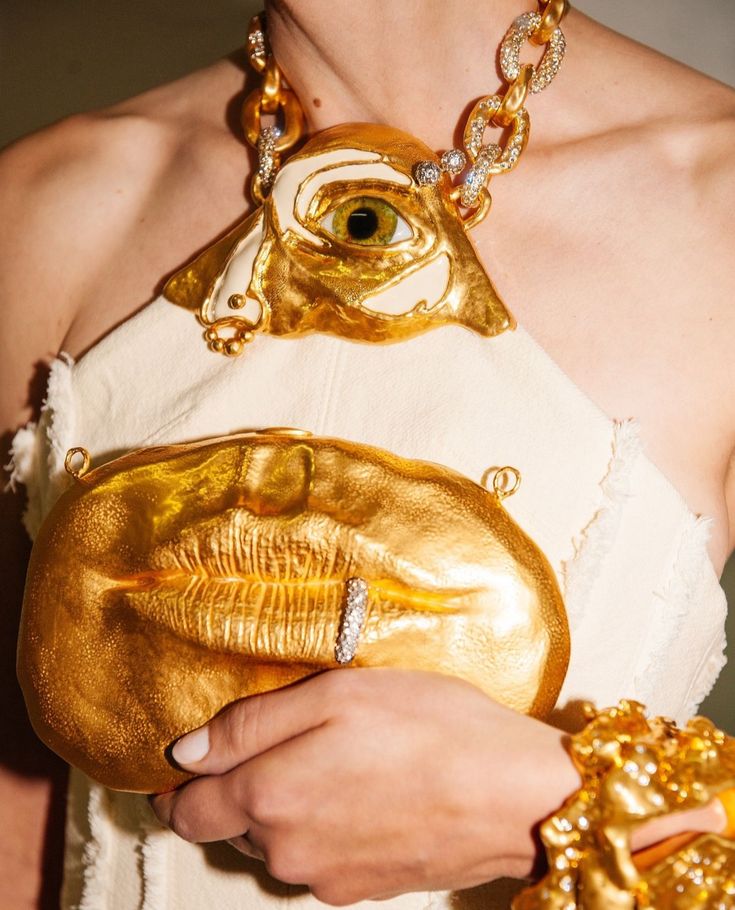 a person is wearing a white strapless top and adorned with unique jewelry. the necklace features a large gold pendant with an eye design. they are holding a gold purse shaped like lips, and a ring is placed near the lips. a gold bracelet is on their wrist.