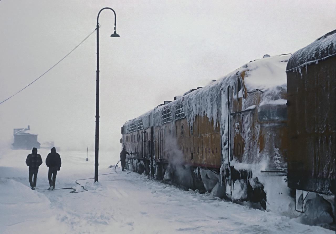 a train covered in snow and ice sits in a snowy landscape with two people walking on the tracks. the train is yellow with brown accents, and the snow covers the top and sides. the train is long, with multiple cars. the two people walking on the tracks are both wearing winter coats and are walking towards the left side of the image. the background is white, and the snow is piled high. the people are walking on the tracks in the middle of the image. there is a streetlight to the left of the image.