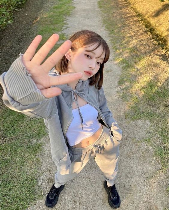 a young woman in a gray hoodie and matching sweatpants stands on a dirt path surrounded by greenery, striking a pose with her hand extended toward the camera. she wears a white crop top beneath the hoodie and black sneakers. the sunlight casts a warm glow on her, creating a natural and vibrant outdoor portrait.  she has short brown hair styled in a modern, tousled look.