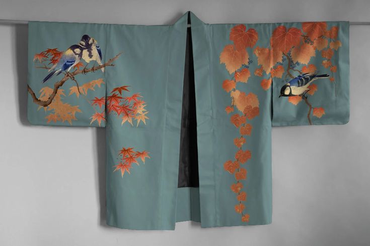 a teal kimono with embroidered designs of birds and leaves is displayed. on the left side are two birds perched on a branch amongst autumncolored maple leaves. on the right side, a bird is also perched on a branch amongst a vine with orange leaves. the kimono is hanging open, revealing a dark interior lining.
