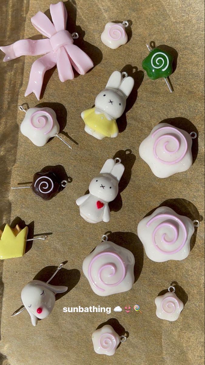 a variety of charms and embellishments are arranged on a flat surface. the objects include a pink bow, several swirl charms in pink and white, a bunny charm in white with a yellow dress, a green lollipop charm, a chocolate lollipop charm, a bunny charm with a red heart, a crown charm, and a charm of a bunny head with a dot for a mouth. the arrangement appears random, with the items scattered across the surface.