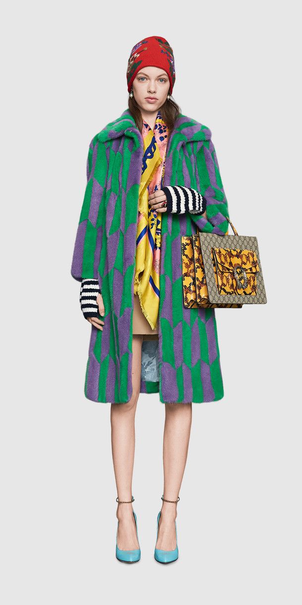 a full shot of a female fashion model posing against a white background. she is wearing a green and purple striped fur coat, a red beanie, a yellow and pink scarf, striped gloves, light blue high heels, and a gucci handbag.