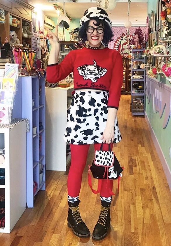 a woman is standing in a vintage store wearing a dalmatian print outfit. she has on a red sweater with a dalmatian on it, a dalmatian print skirt, red tights, and black boots. she is also wearing a dalmatian print hat, a choker, and glasses. she is holding up a peace sign with her left hand and has a dalmatian print handbag hanging from her right hand. the store is filled with vintage items.