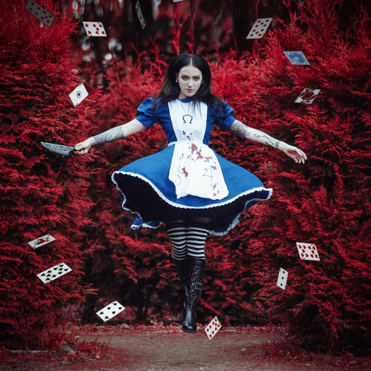 the image depicts a young woman cosplaying as a dark, gothic interpretation of alice from alice in wonderland. she's airborne, midleap, dressed in a blue and white dress with blood splatters, an apron bearing an omega symbol, and striped stockings. she holds a knife in one hand.  playing cards are scattered around her, falling from the sky, against a backdrop of crimsonhued foliage. the overall atmosphere is dark, surreal, and slightly macabre, suggesting a darker, more sinister take on the classic tale. the highquality photography enhances the gothic and fantasy elements.