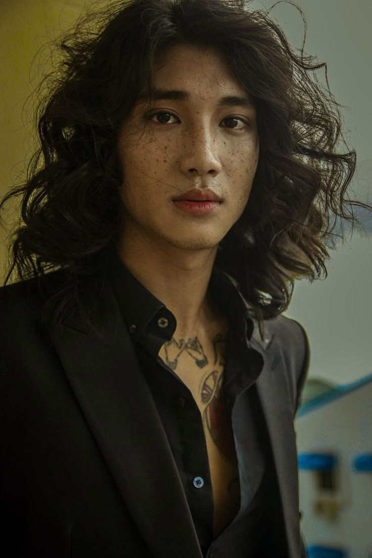 a portrait of a man with freckles and long wavy hair, wearing a black suit over an open shirt, revealing a tattoo on his chest.