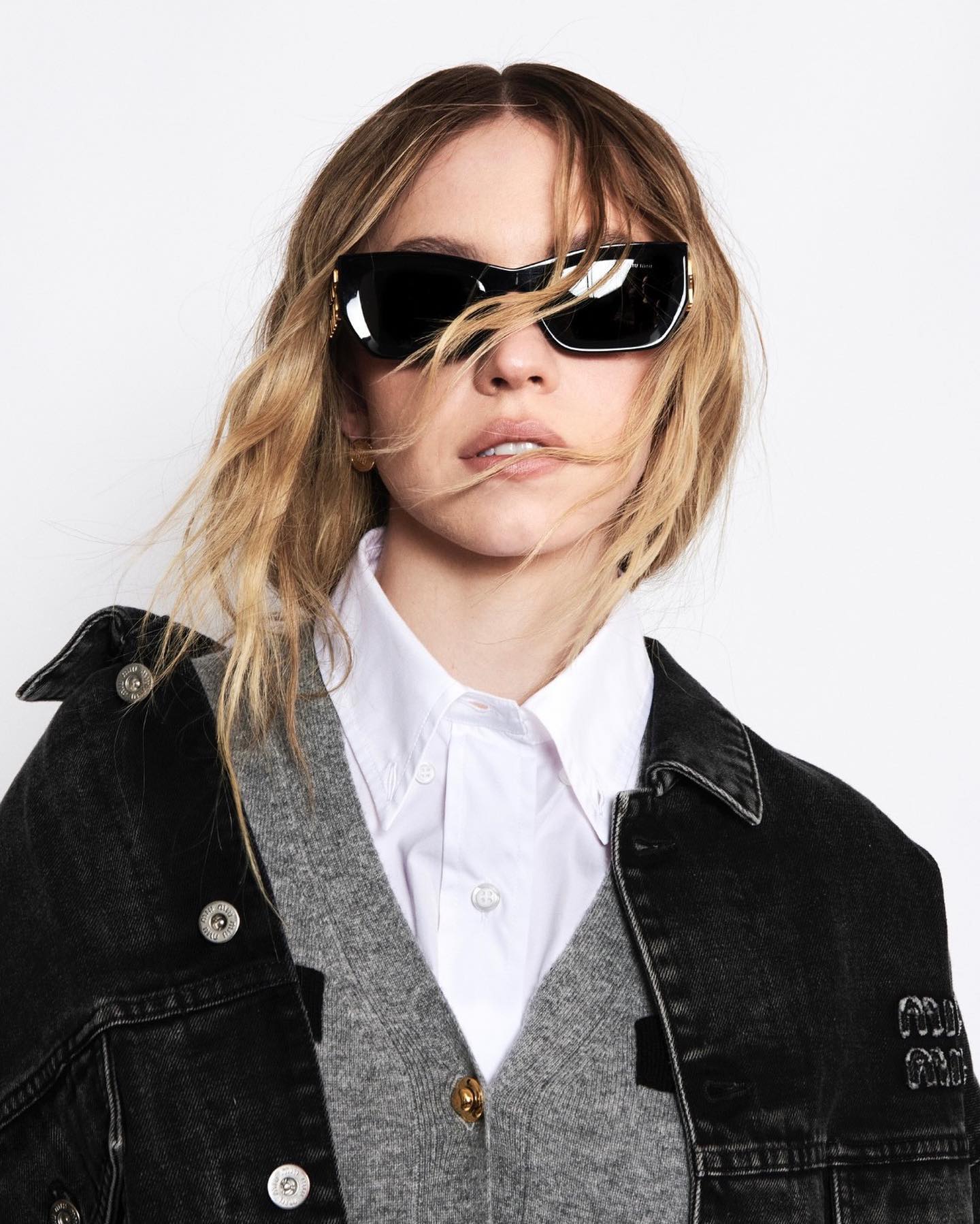 a closeup portrait of a blonde woman modeling a stylish outfit.  she is wearing black cateye sunglasses, a grey cardigan over a white buttondown shirt, and a black washed denim jacket. her hair is styled in loose waves. the background is a plain white, making the clothing and accessories the focal point. the overall style is casual yet chic.