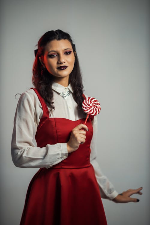 a young woman with dark hair and dark makeup is dressed in a red dress over a white shirt. she is holding a red and white lollipop and looking directly at the camera. the background is plain gray.