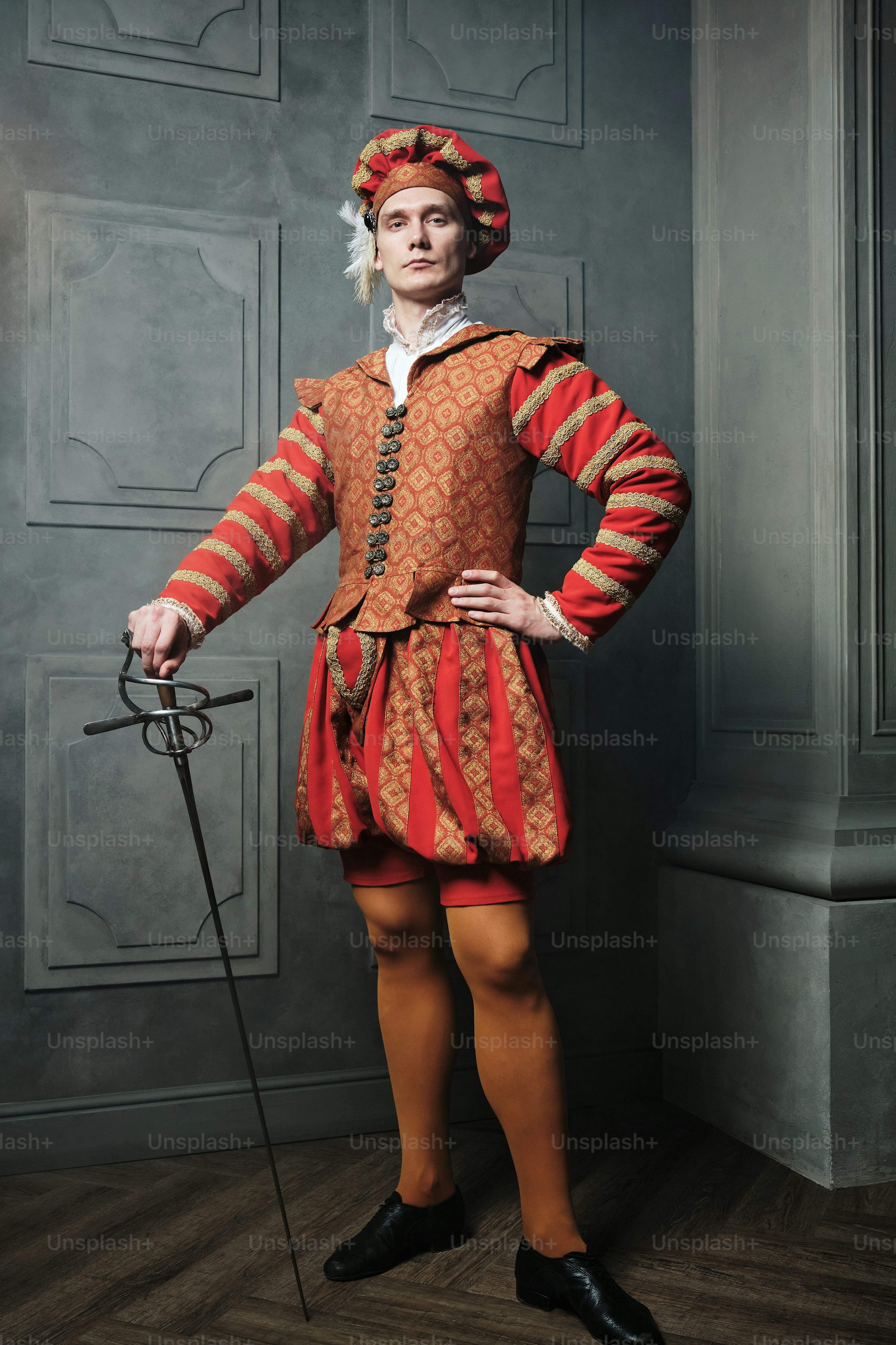 a fulllength portrait of a man dressed in a vibrant red and gold renaissanceera costume. he stands confidently, holding a rapier, in a richly detailed setting.  his clothing is intricately patterned and includes a doublet, breeches, and a striking hat with a feather. the overall image evokes a sense of historical grandeur and elegance.