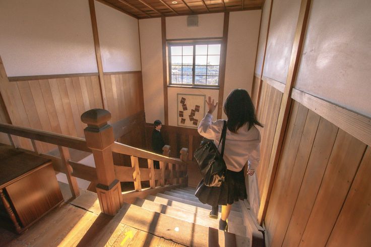 the image shows a japanese schoolgirl in a uniform, wearing a backpack, ascending a wooden staircase in an old, traditional japanese building.  she's waving her hand, possibly in farewell or greeting. sunlight streams in through a high window illuminating the wooden walls and floors of the hallway. another student is visible further up the stairs. the overall aesthetic is nostalgic and reminiscent of a classic japanese school setting.