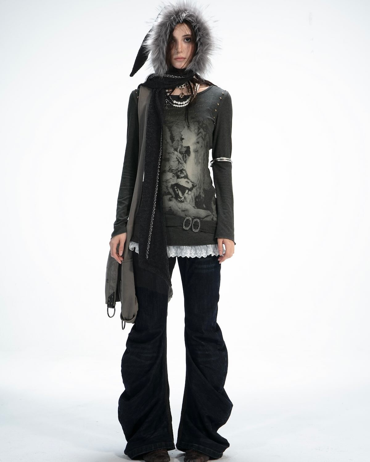 a fullbody studio shot of a young woman showcasing a layered outfit. she's wearing a dark gray longsleeved shirt with a wolf graphic, layered over a delicate lace camisole. the shirt features subtle detailing like studs or stitching at the shoulders.  she's also wearing dark wash wideleg bellbottom jeans and a long, chunky, dark gray scarf draped around her neck and shoulders, which is partially concealed by an oversized gray fur hood. the model is accessorized with several layered necklaces and a bracelet. her pose is relaxed, standing with her arms slightly down. the overall style is a blend of bohemian, grunge, and alternative aesthetics. the background is a plain white, highlighting the clothing and the model.