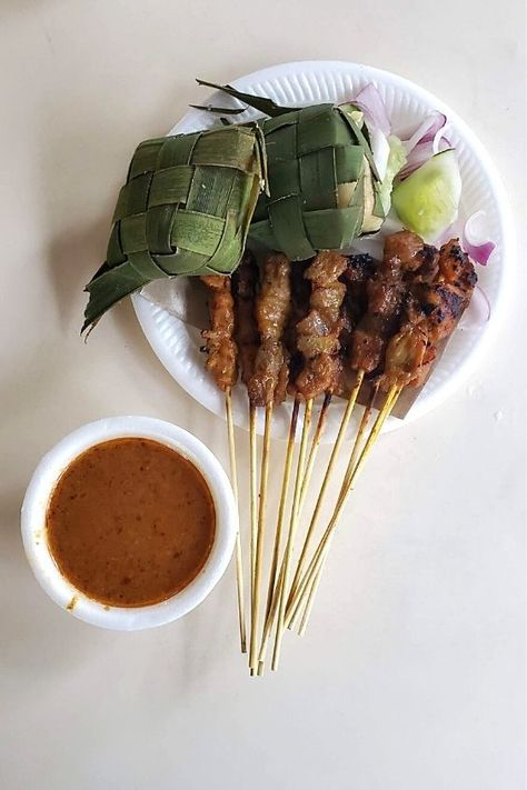 a topdown view of a plate of satay, accompanied by a small bowl of peanut sauce and two ketupats (diamondshaped rice cakes wrapped in palm leaves). the satay consists of grilled meat on bamboo skewers and is arranged attractively on the plate along with sliced onions and cucumbers. the scene is set on a light beige surface, suggesting a casual dining setting. the image is welllit and captures the vibrant colors and textures of the food.