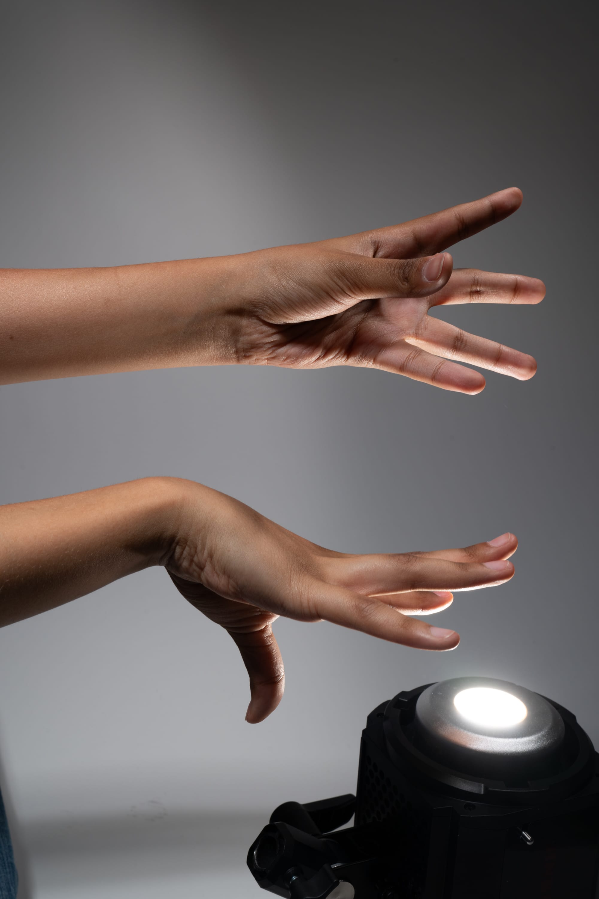 the image shows two hands interacting with a spotlight in a studio setting. the top hand is open, as if gesturing towards the light, while the lower hand is positioned below, fingers outstretched as if feeling the light's warmth. the spotlight is positioned in the lower right corner of the image, casting a bright white light.  the background is a gradient of grays, creating a sense of depth and highlighting the hands and light source.