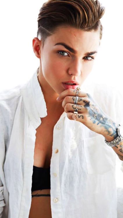 a portrait of ruby rose, a woman with short, styled brown hair and an undercut. she is wearing a loosely buttoned white shirt, revealing a black bra. she has tattoos on her hand and arm, and multiple rings on her fingers. she is looking at the camera with a slight smirk, her hand held up to her mouth in a contemplative pose. the background is light and neutral.