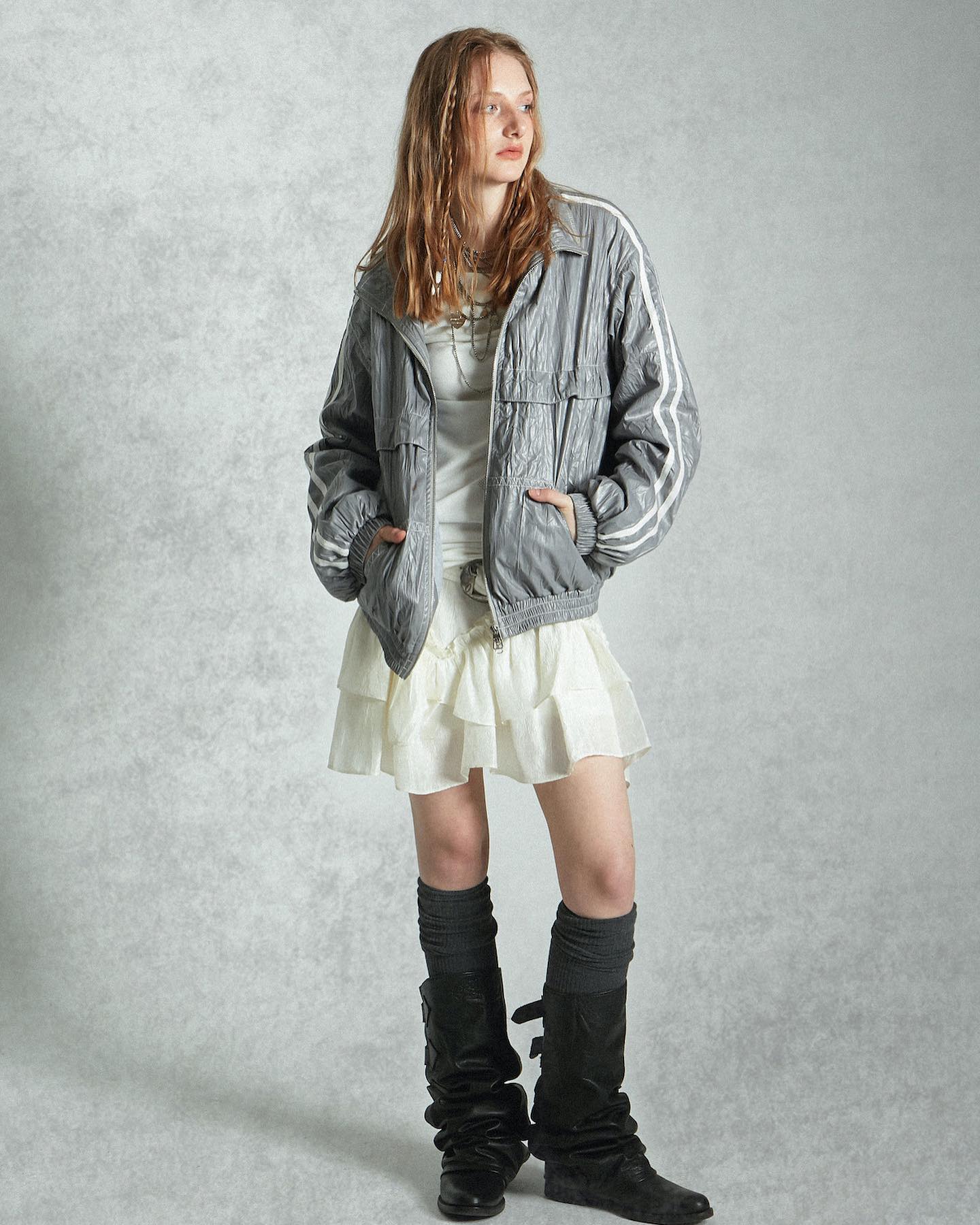 a fulllength shot of a woman in stylish clothing poses against a neutral background. she's wearing a grey jacket with white stripes, a white ruffled miniskirt, and long black boots with dark grey socks.  the jacket is slightly oversized and has a relaxed fit. the skirt is layered and adds a touch of femininity.  her hair is styled loose and wavy, framing her face. the overall look is casual yet fashionable.