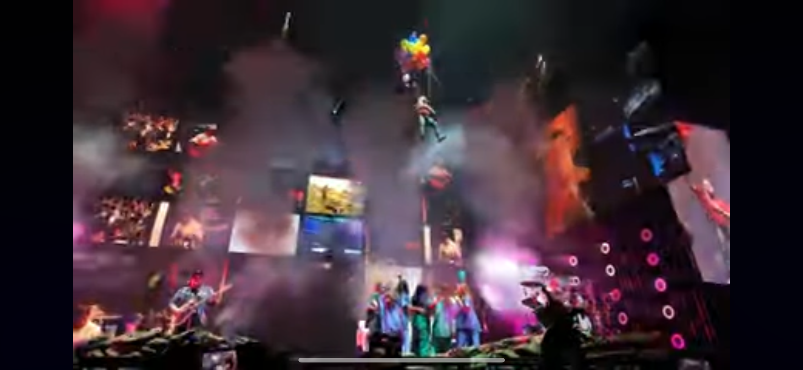 this is a vibrant, energetic shot from a concert. the stage is brightly lit with spotlights, and a band is performing for a large, enthusiastic crowd. there are balloons floating high above the stage, adding to the festive atmosphere. the photo captures the excitement and energy of a live concert, with the band members passionately playing their instruments and the audience cheering them on. the stage is adorned with various screens, showcasing different images and videos, adding to the visual spectacle of the event. this image perfectly encapsulates the excitement and joy of a live music experience.