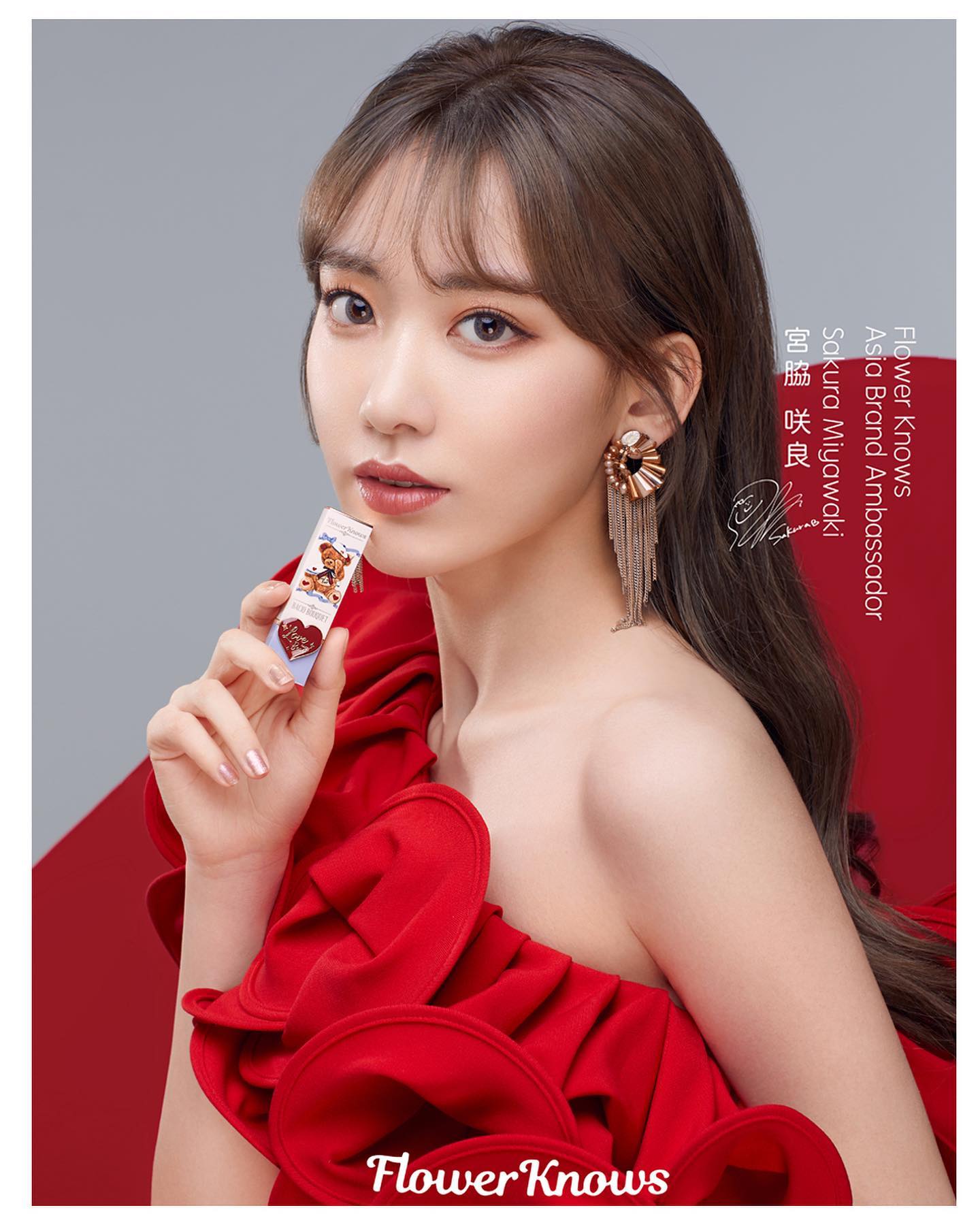 a woman in a red dress with a red flower design is holding a lipstick up to her lips. the lipstick is a light pink color and has a picture of a bear on it. the woman has long brown hair and is wearing red lipstick. the background is white. the text "flower knows" is written in pink at the bottom of the image. the text "flower knows asia brand ambassador" is written in white above the woman's head. the text "sakura miyawaki" is written in white below the woman's chin.