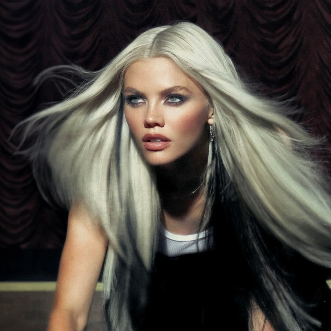 a portrait of a woman with long, whiteblonde hair and dark roots. she has smoky eye makeup and wears a white tank top. the background is a dark, textured brown. she has silver earrings.