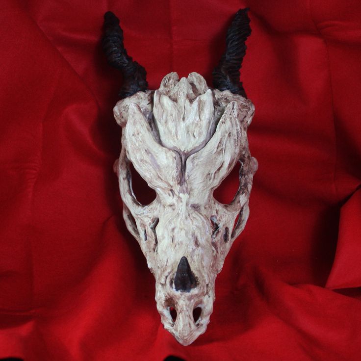 a closeup shot of a white dragon skull mask with black horns. the mask is made of a textured material that resembles bone. the mask has large, empty eye sockets and a gaping maw with sharp teeth. it is set against a backdrop of red fabric, creating a stark contrast.