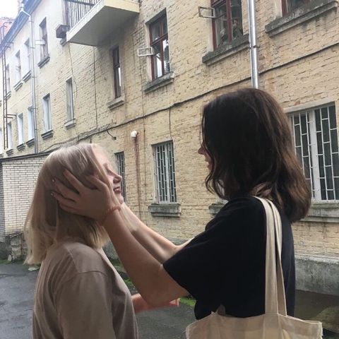 two young girls, one with blond hair and one with dark hair, stand in an alleyway outside a building. the blond girl's hand rests on her friend's head and she is looking up at her friend. they are both smiling. the image is in focus and captures the moment of warmth and happiness between the two friends.