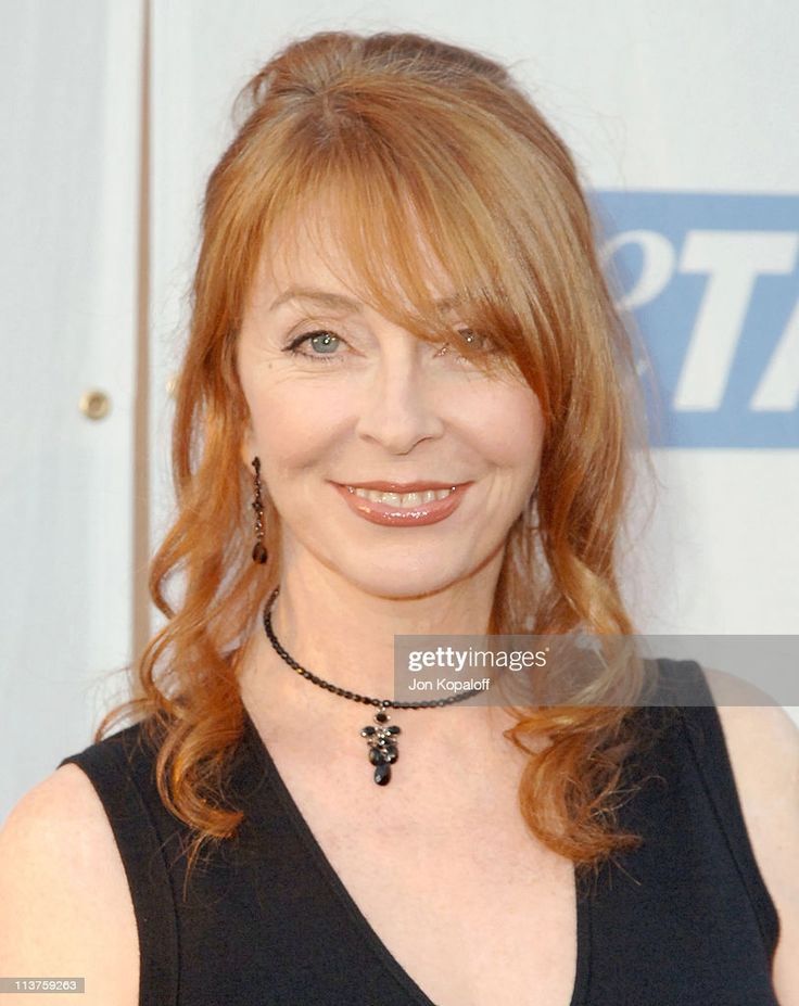 a woman with long red hair smiles for the camera. she is wearing a black dress with a necklace and earrings. she has a white background and is looking at the camera.