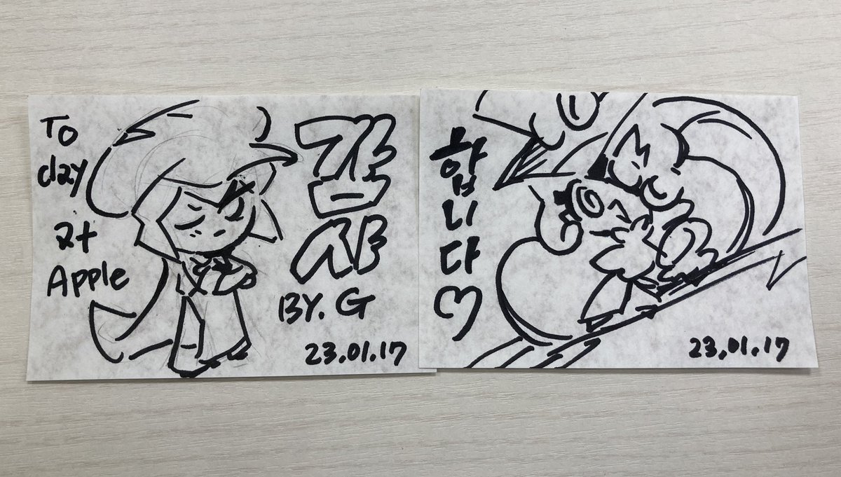 the image is of two pieces of paper placed side by side on a wooden table. on each piece of paper is a black ink cartoon drawing, an autograph, and a date. the drawings are cartoons, one of a humanoid character and the other of two abstract figures riding a wave. the table top is light brown wood grain.