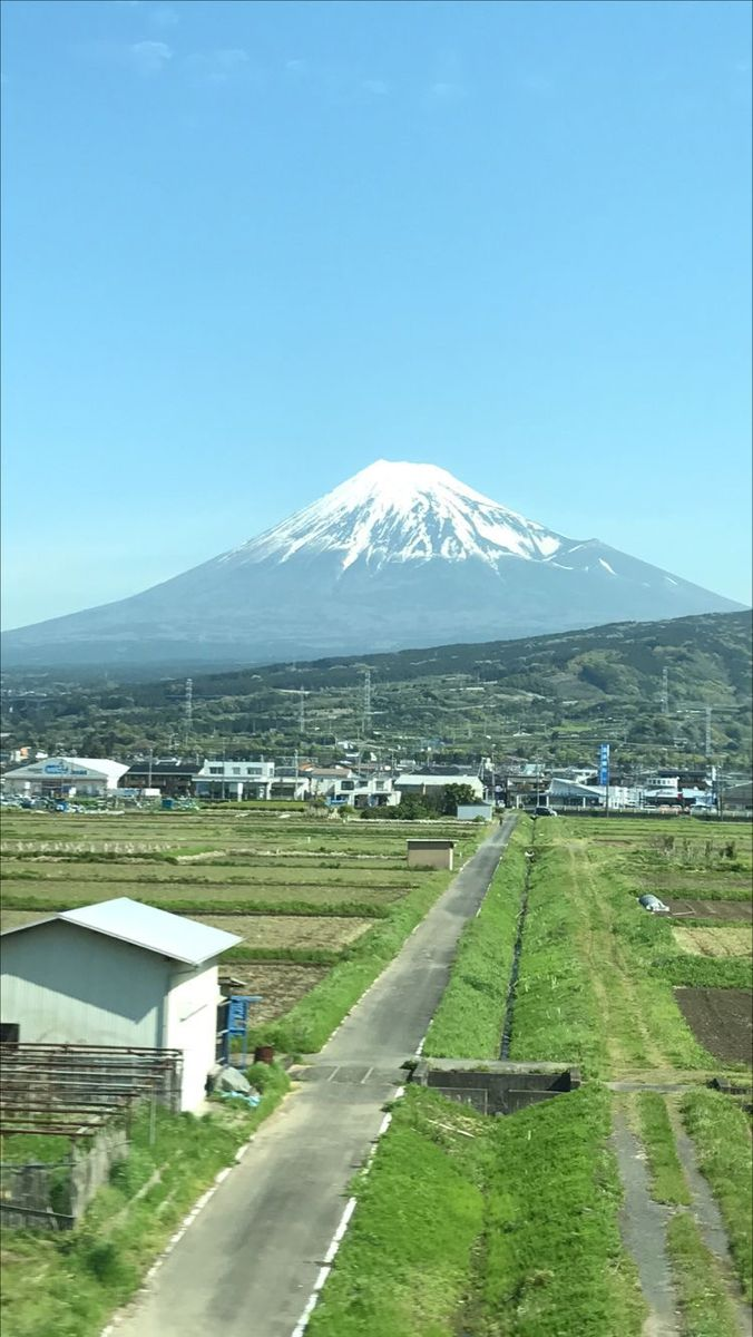 a scenic view of mount fuji under a clear blue sky, with a rural landscape featuring rice paddies and a narrow road leading towards the mountain. a building stands to the left of the road, adding to the rural setting. the mountain's peak is covered in snow, contrasting with the greenery below.