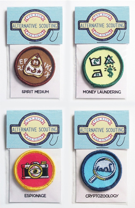 the image shows four sets of alternative scouting badges. each set includes a header card that reads "alternative scouting for boys and girls," and a smaller circular embroidered patch.  the patches depict: a ouija board (spirit medium), money laundering symbols, a camera with an eye (espionage), and a magnifying glass over a creature (cryptozoology). the patches are displayed on a white background, each in its own clear plastic sleeve.  the style is whimsical and slightly subversive.
