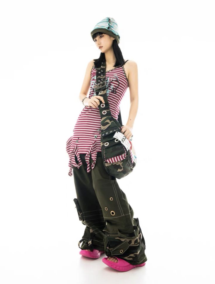 a fulllength studio shot of a young woman model posing against a white background. she's wearing a pink and white striped sleeveless dress, olive green camouflage pants with various pockets and straps, a light teal beanie, and bright pink sandals. she's carrying a camouflage shoulder bag. the overall style is casual yet trendy, featuring a mix of textures and patterns.  the image is welllit and focuses on the model's outfit and pose.