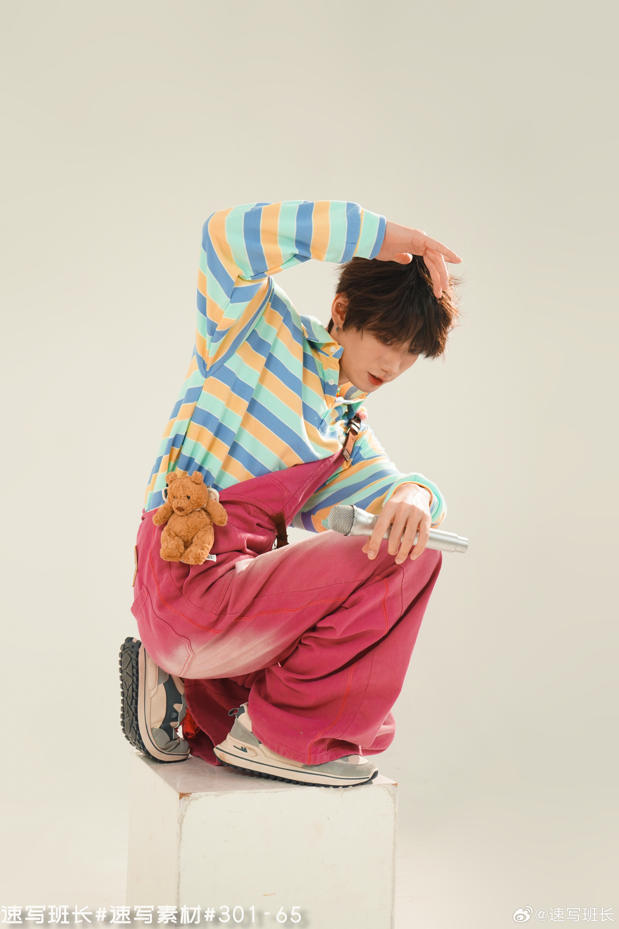 a young man with dark hair is posing for a photo shoot in a studio. he is wearing a colorful striped shirt, pink overalls, and sneakers. he has a teddy bear attached to his overalls and is holding a microphone in his hand. he is striking a playful pose while looking down and shielding his eyes with his other hand. the backdrop is a plain offwhite color. the image is welllit with studio lighting.