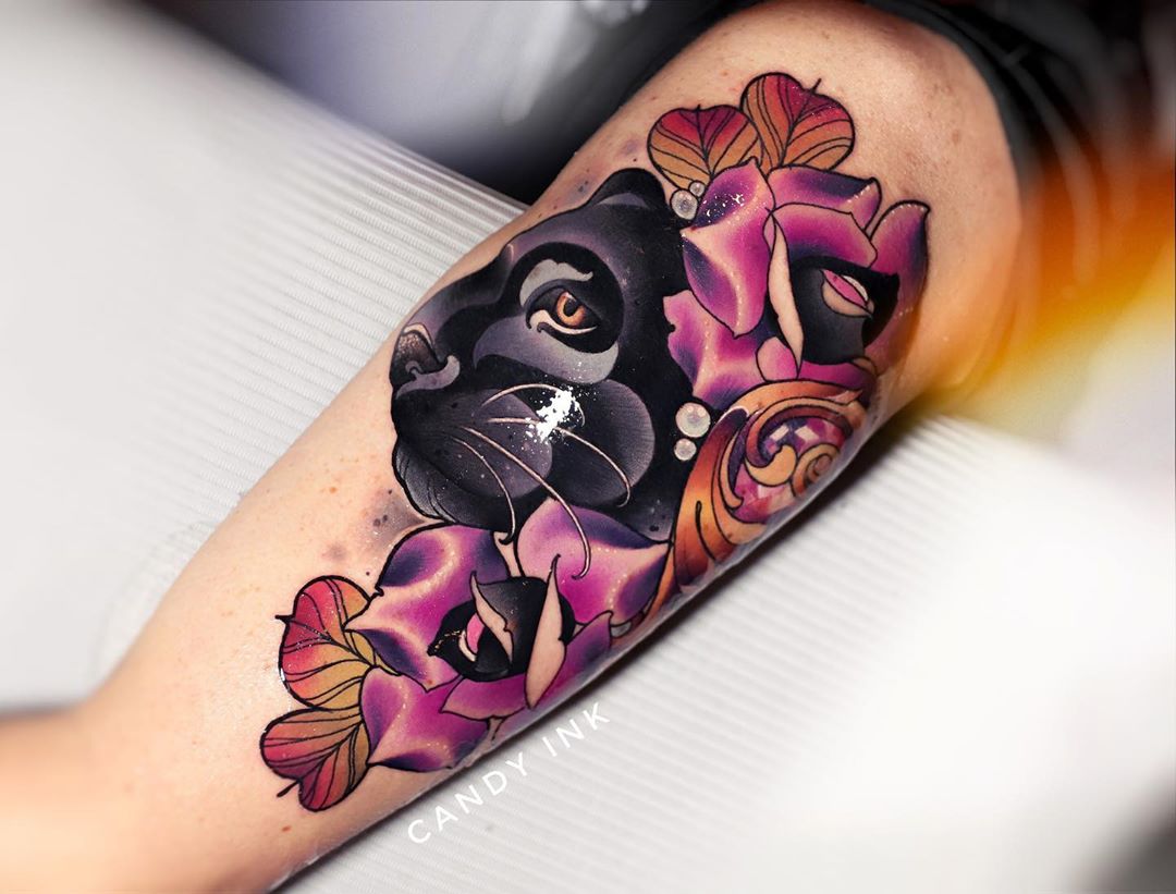 a closeup shot of a newlydone neotraditional black panther tattoo surrounded by colorful flowers on someone's inner forearm. the tattoo has bold lines and is done in a variety of colors, including black, purple, red, and yellow. the tattoo is set against a white background.