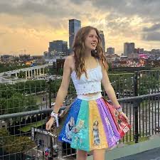 a young woman in a colorful rainbow skirt and white top stands on a city street, smiling and looking at the camera.  she is surrounded by buildings. she is wearing a colorful rainbow skirt with images on it, and a white top with a lace trim. her hair is brown and pulled back off her face. she is smiling and looking directly at the camera. the background is an urban cityscape.