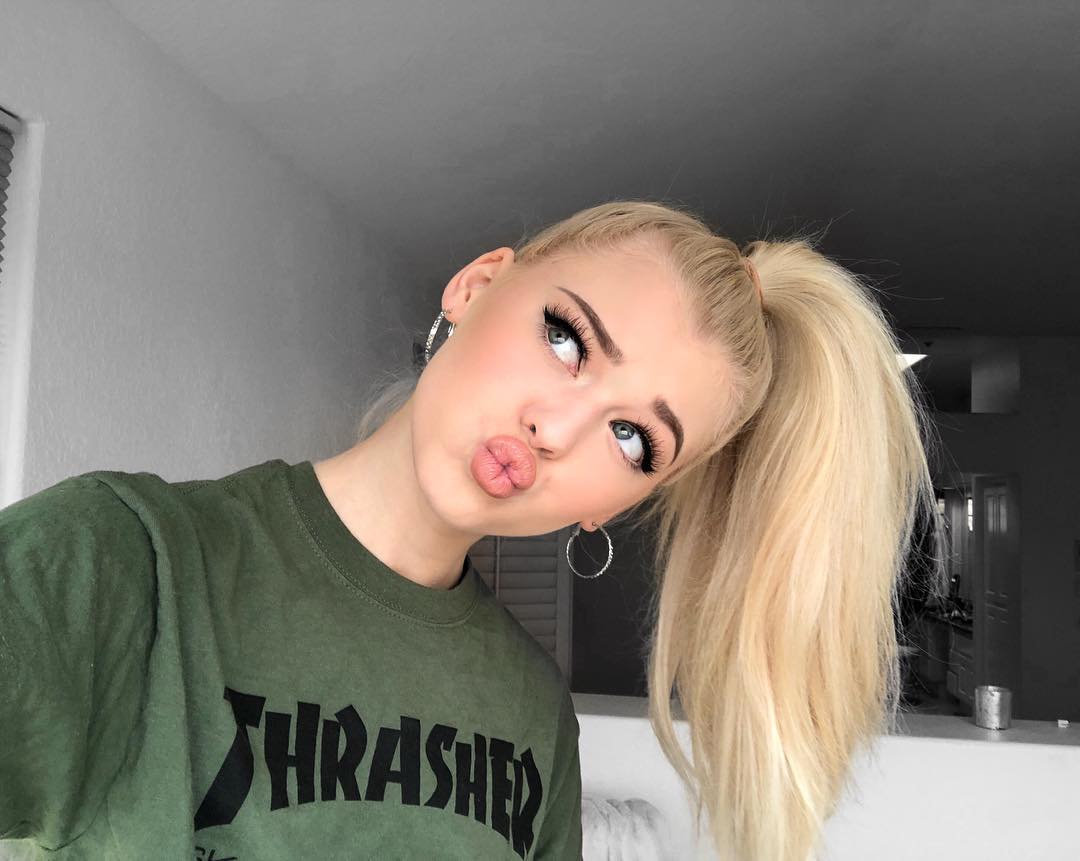 a young woman with blonde hair in a high ponytail is taking a selfie. she is pouting her lips and looking up. she is wearing a green thrasher tshirt and hoop earrings. the background is a white wall and a doorway.