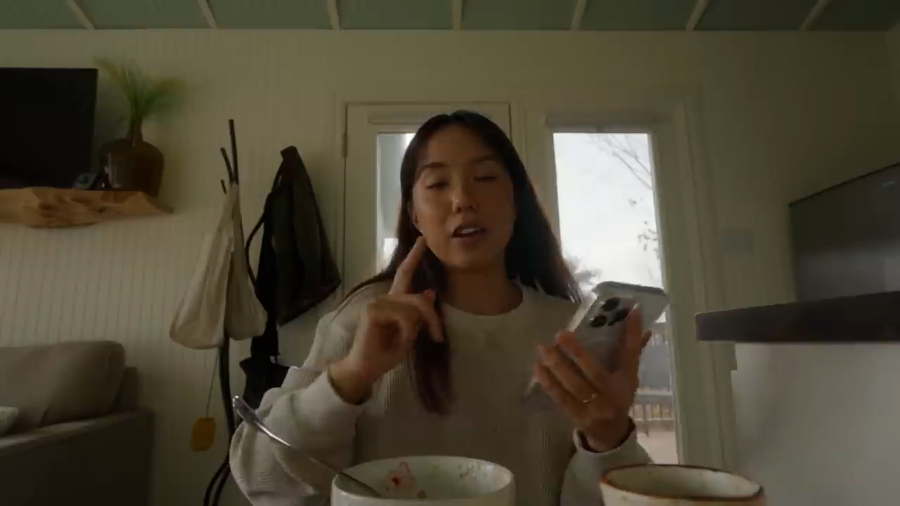 a young asian woman sits in the kitchen of her tiny home, talking to her phone while eating breakfast.  the space is bright and airy, with white walls and natural wood accents. the image has a warm and inviting feel.