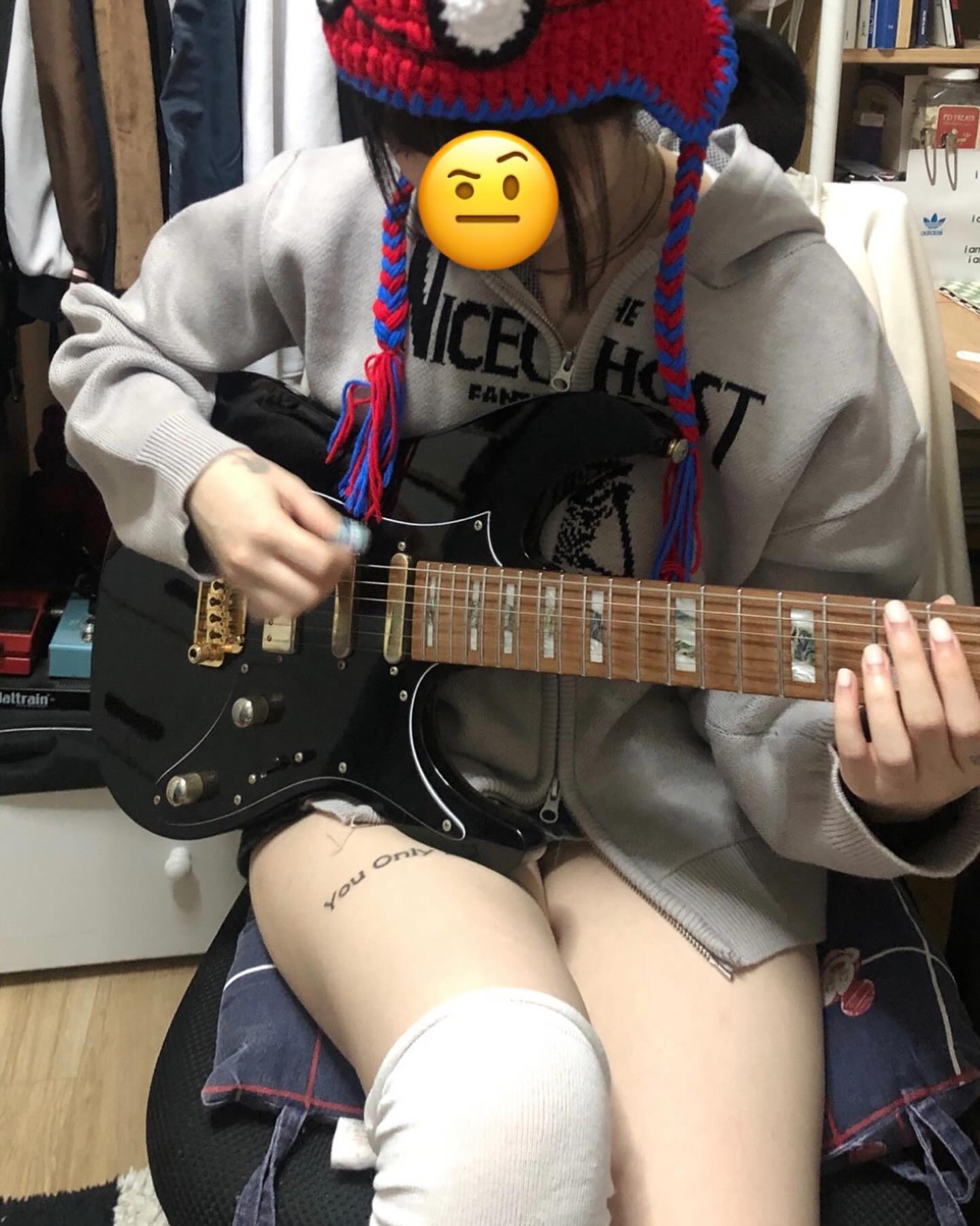 a young woman sits on her bed in her bedroom, holding a black electric guitar. she is wearing a gray hoodie, white kneehigh socks, and a red, white, and blue beanie. her face is covered by a digital emoji. the guitar is black with gold hardware. the room is cluttered, and the walls are white. the lighting is artificial and comes from the ceiling.