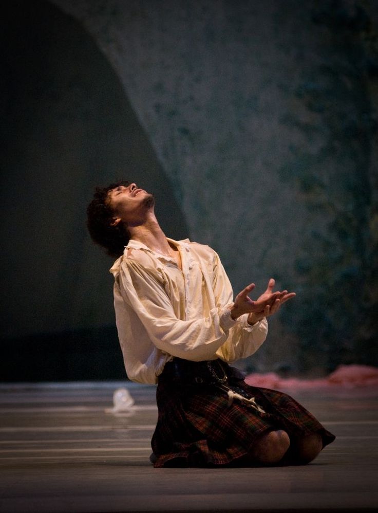 a male ballet dancer wearing a white shirt and kilt is kneeling on stage with his arms outstretched and head tilted upwards, he is passionately expressing emotions in this powerful and dramatic moment in his performance. the background is a dark, shadowy theater stage with lighting focused on the dancer.
