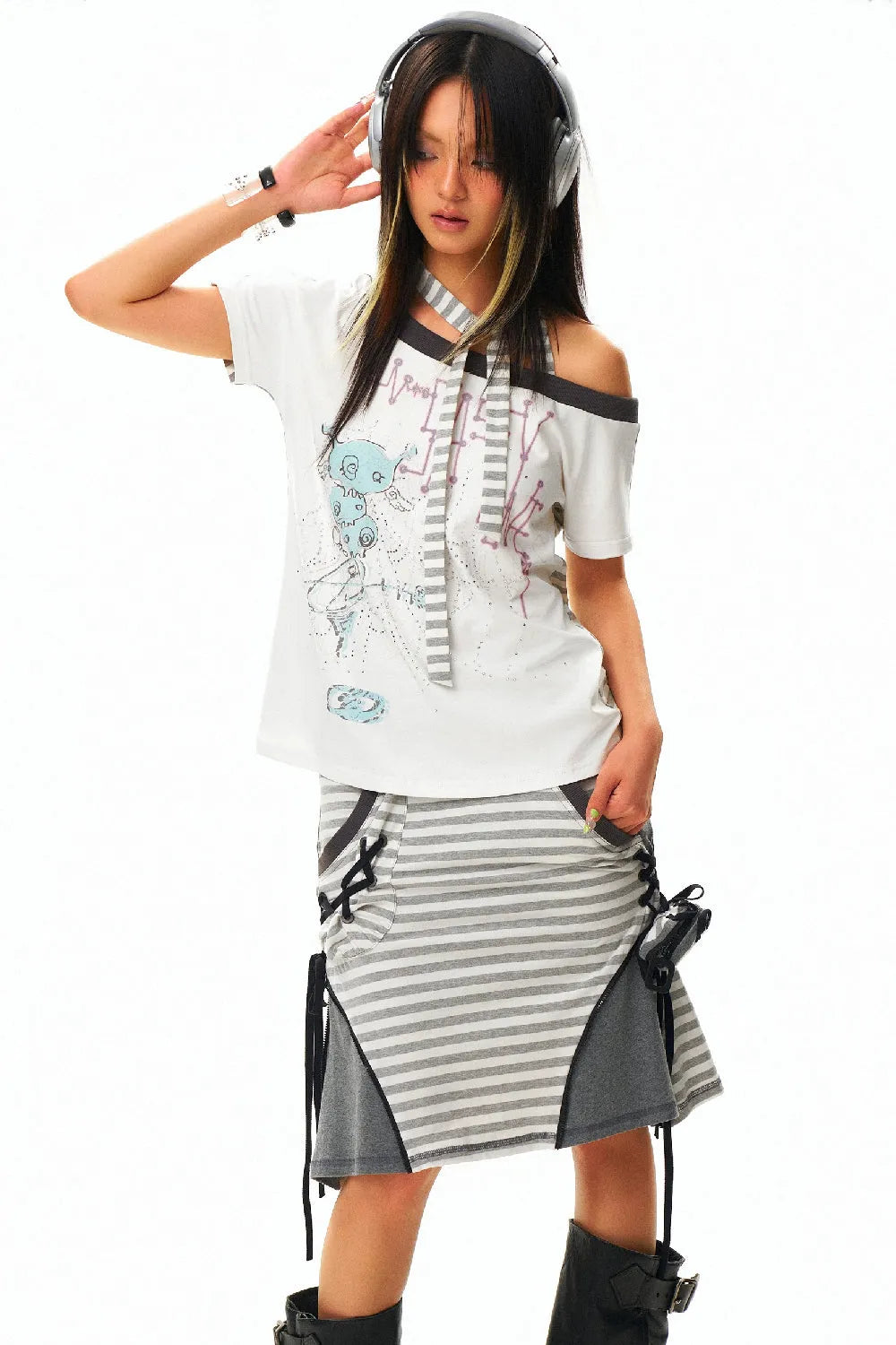 young asian woman, possibly a model, is posing in a studio against a plain white background. she is wearing a stylish outfit that includes a white offtheshoulder graphic tee and a grey and white striped skirt with a grey panel detail. she also has on black boots. a pair of headphones rest on her head, and she has several bracelets on her left wrist, suggesting she may be listening to music. her hair is styled in a way that partially obscures her face. the overall aesthetic is casual and trendy.