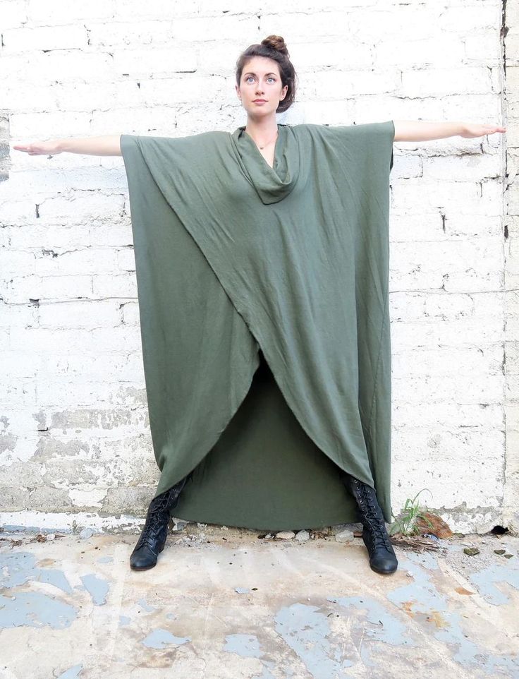 a woman stands in front of a white brick wall with her arms outstretched. she is wearing a long, olive green poncho with a cowl neck. the poncho has an open front, revealing her legs and black laceup boots. her hair is pulled back in a bun.