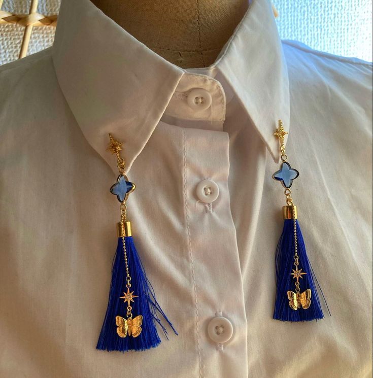 closeup view of a white collared shirt adorned with two identical collar pins. each pin features a gold chain with a blue crystal clover, a blue tassel, a gold star charm, and a gold butterfly charm hanging from it. the shirt and the jewelry are neatly presented, with the collar pins symmetrically placed on either side of the shirt's center. the background is slightly blurred, suggesting an indoor setting. the overall aesthetic is elegant and sophisticated.