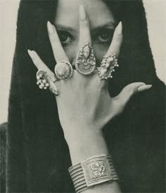 a black and white portrait of a woman wearing a variety of rings and a bracelet on her hand. she is wearing a dark, flowing garment and her expression is mysterious and alluring. the image is evocative of the 1960s and 1970s bohemian style, and the woman appears to be a free spirit. the jewelry is ornate and detailed, suggesting a taste for the exotic and mystical. the woman's beauty is enhanced by her dark hair and makeup, and she embodies the allure of the bohemian lifestyle.