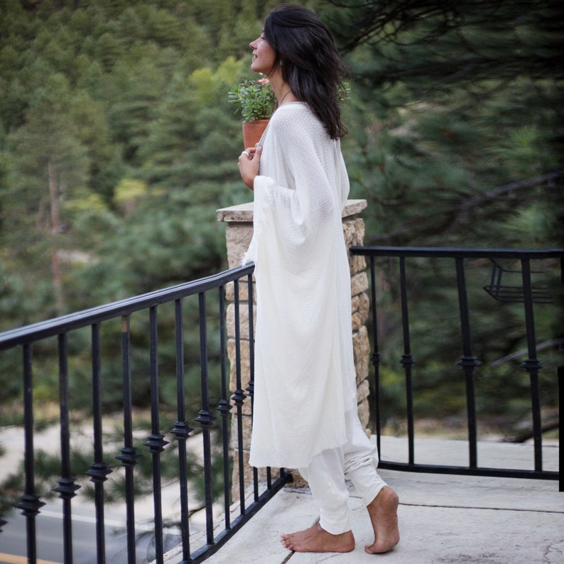 a woman in a long, white robe stands on a balcony overlooking a mountainous landscape. she holds a small pot of plants, and her gaze is directed towards the view. the scene is peaceful and serene, suggesting a moment of calm and reflection. the woman's clothing and posture suggest a relaxed and spiritual state, possibly connected to meditation or yoga. the natural setting adds to the sense of tranquility and wellbeing.