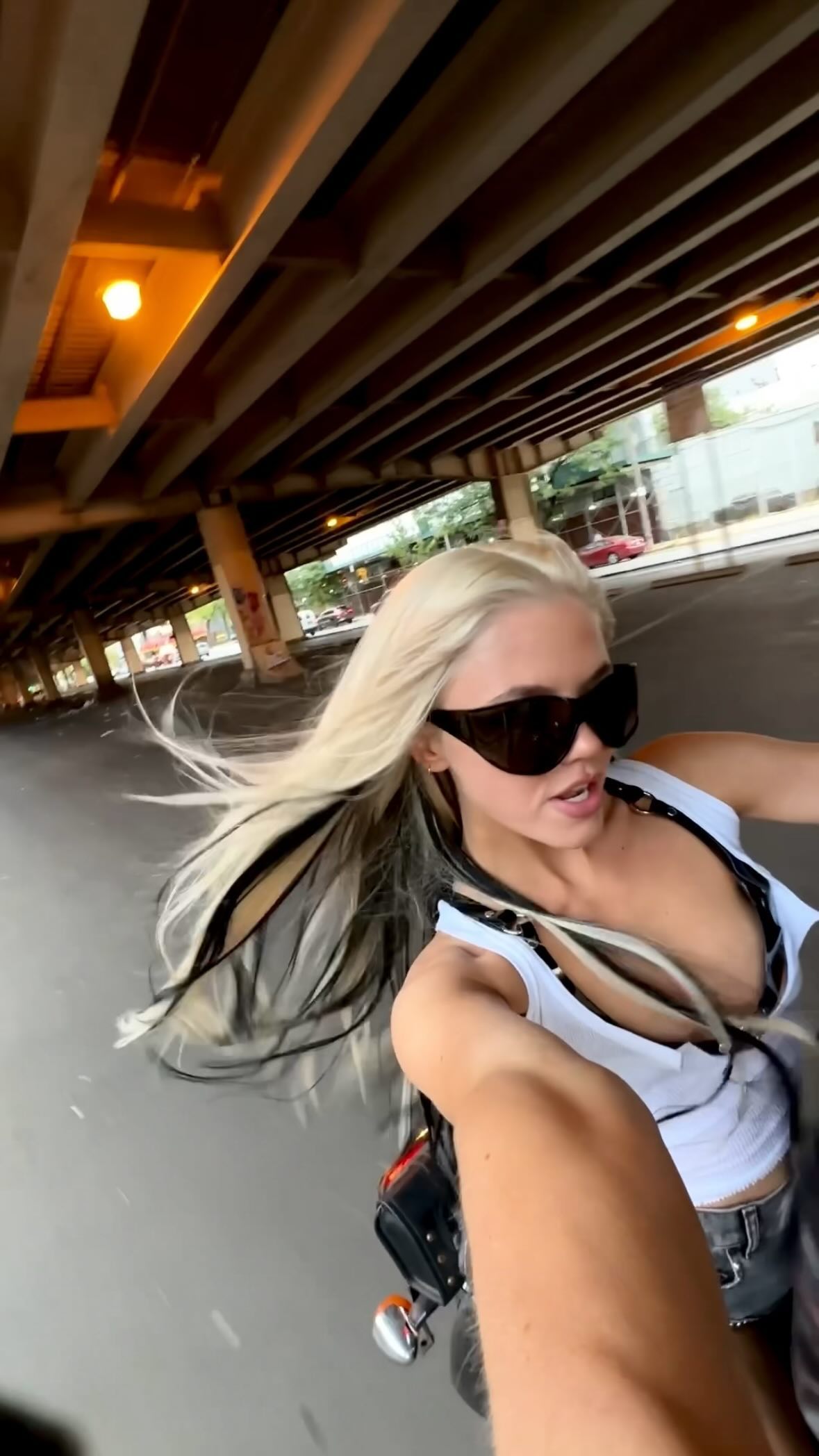 a woman with blonde hair and black streaks is riding a motorcycle down a street, underneath an overpass. she is wearing sunglasses and a white tank top with significant cleavage. the background shows urban buildings, traffic, and light fixtures under the overpass. the scene takes place during the daytime.