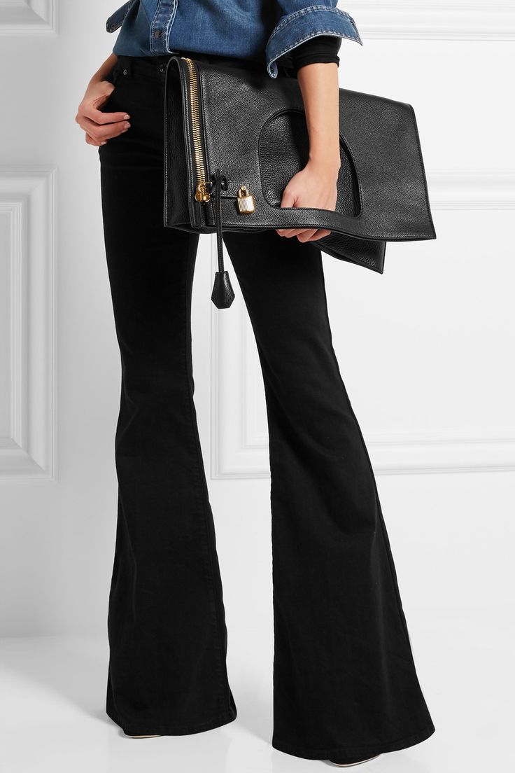 a person is shown from the midtorso down, wearing black flare pants and a denim shirt. they are holding a large black leather handbag with gold hardware and a padlock detail. the background is a plain white wall.