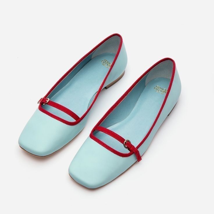 a pair of light blue leather flats with a red strap across the vamp are presented against a white background.  the shoes are positioned slightly angled, with one shoe slightly overlapping the other. the brand name is subtly visible on the insole. the shoes appear to be designed for women.
