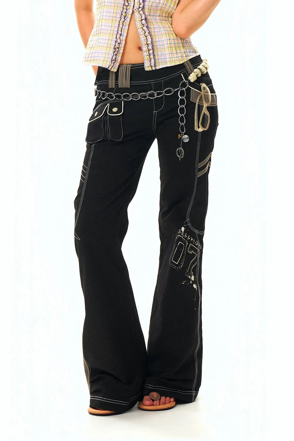 a fullbody studio shot of a young woman standing against a white background, wearing stylish black bell bottom jeans adorned with embellishments, a chain belt, and sandals.  she's also wearing a light colored plaid shirt. the image highlights the details of the clothing and the model's pose.