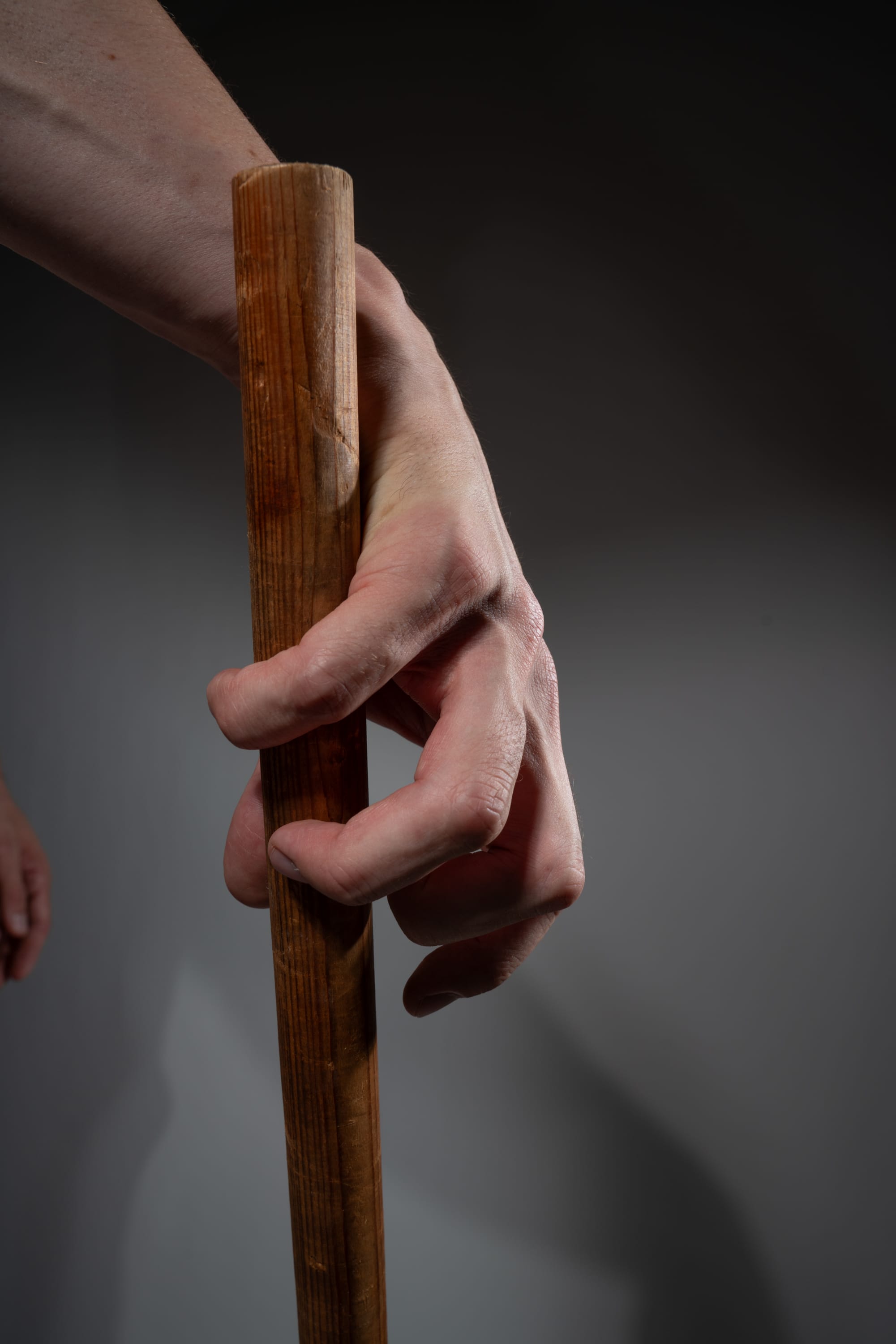 closeup shot of a person's hand tightly gripping a wooden stick against a gray background. the hand is illuminated by soft light, creating subtle shadows and highlighting the texture of both the skin and the wood.