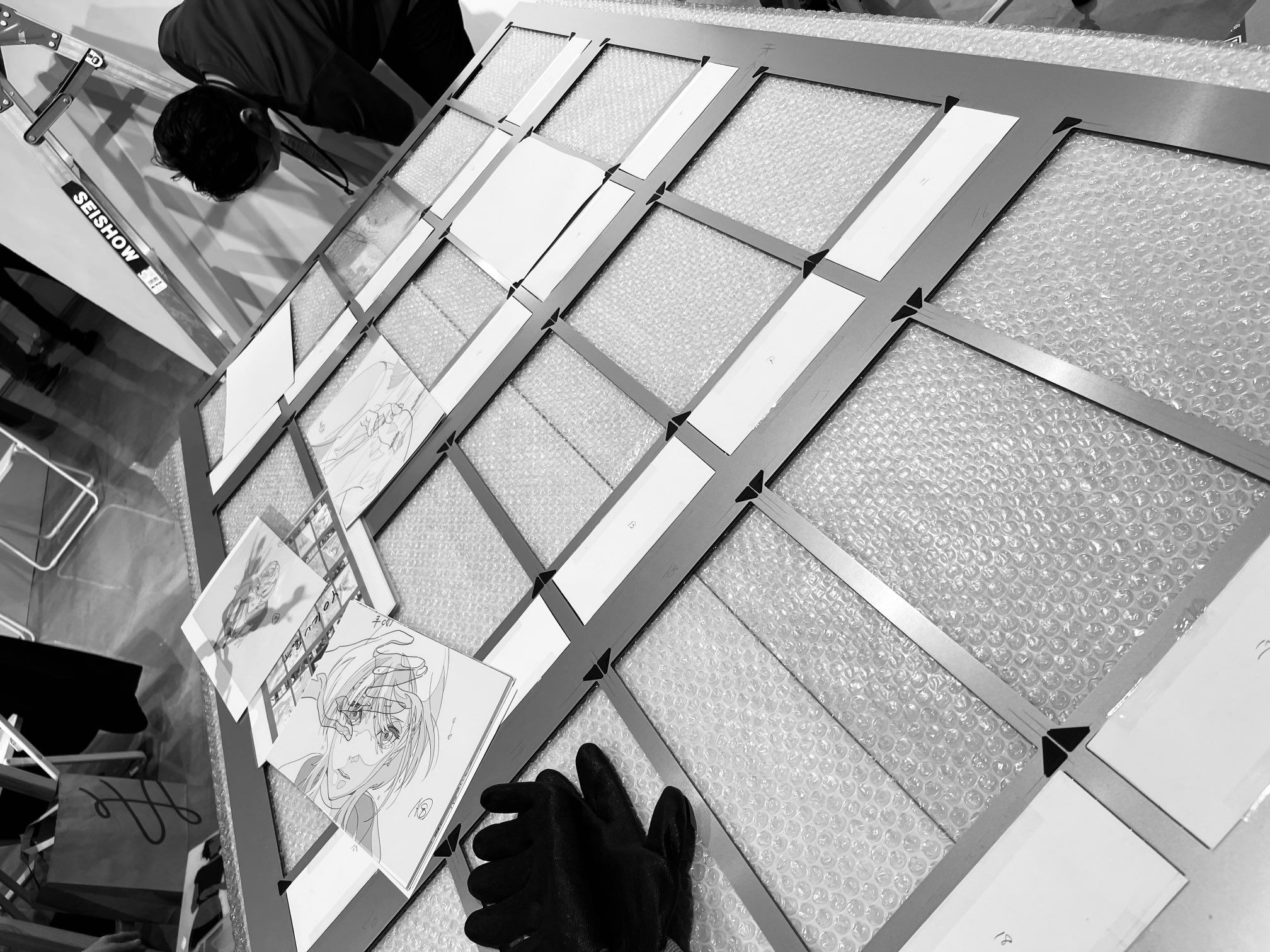 black and white photograph of workers arranging framed manga artwork for an exhibition. bubble wrap protects the floor and potentially the artwork itself. the perspective is from above, offering a behindthescenes glimpse into the preparation.