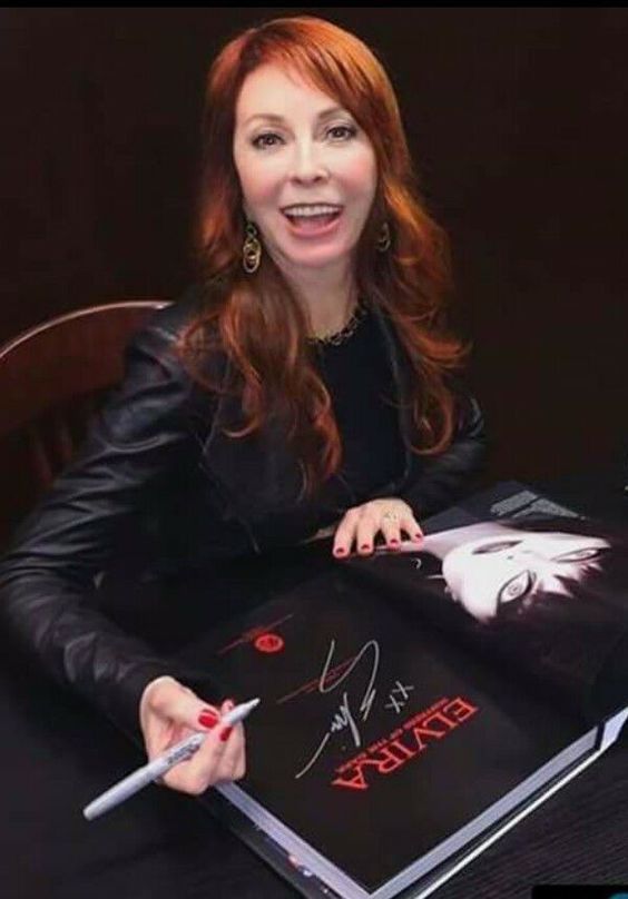 elvira, mistress of the dark, is signing a book. she is sitting at a table and wearing a black leather jacket.  the book is in the foreground, and her autograph is visible.  she is smiling and holding a pen.