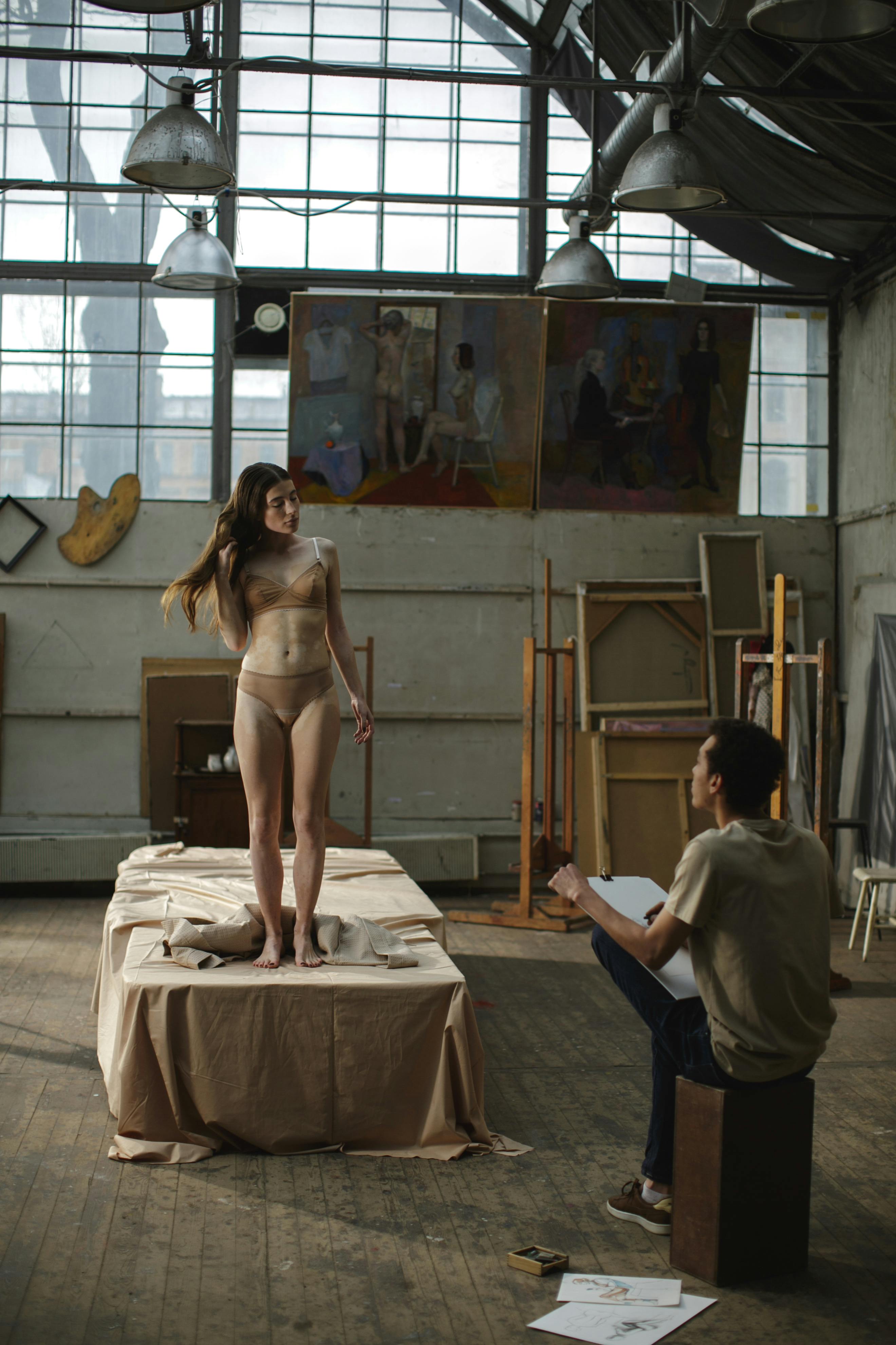 a female model stands on a draped platform in an art studio while a male artist sits and sketches her. the studio has large windows, industrial lighting, and artwork on the walls. the model is wearing a beige bra and underwear. the artist is focused on his work. the overall atmosphere is one of creativity and concentration.